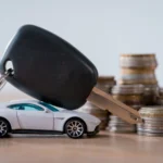 miniature car, car keys and metal coins on wooden surface isolated on grey